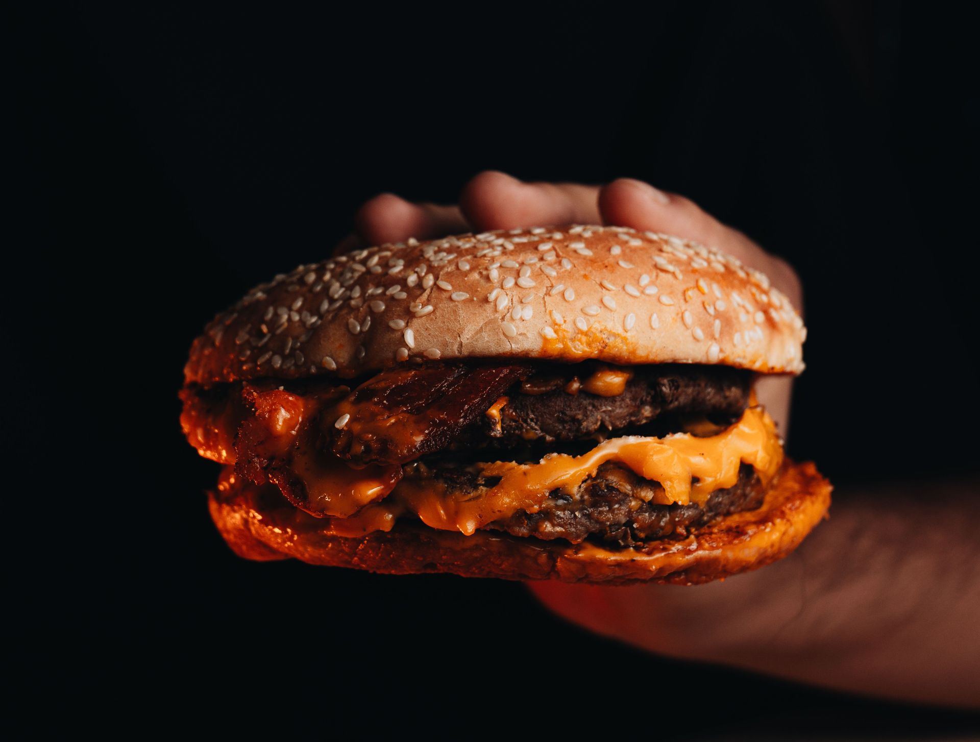 Una mano che regge un cheeseburger ai semi di sesamo su uno sfondo scuro.