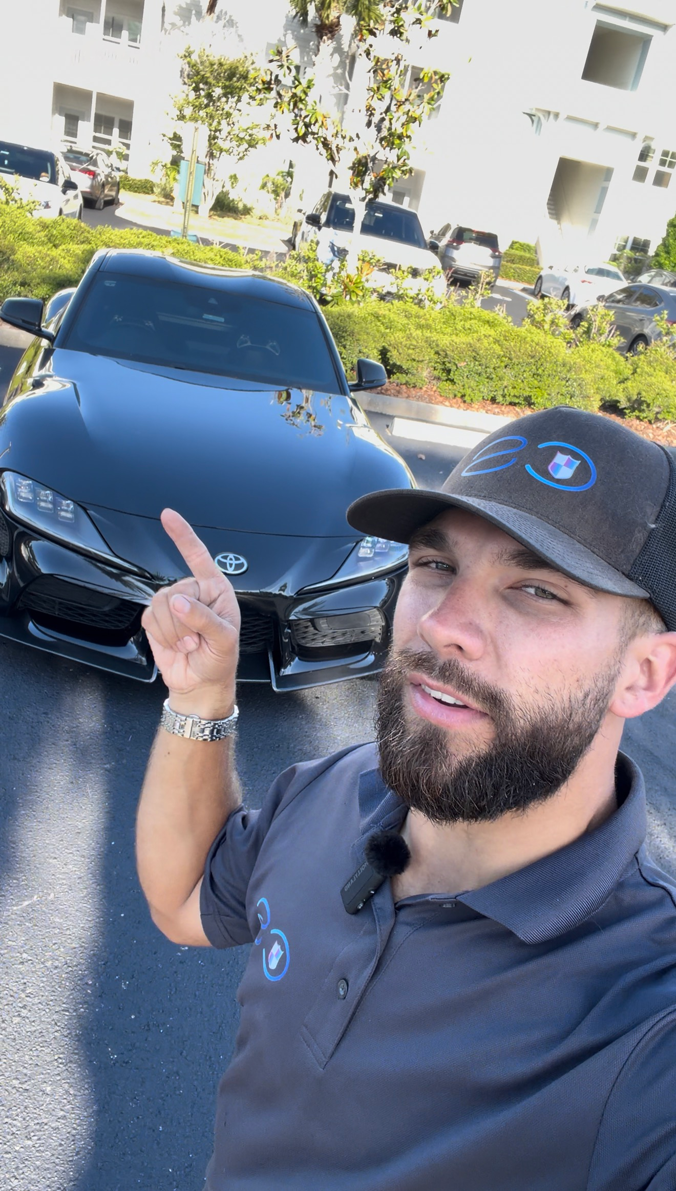 Man taking a selfie and pointing at a black Mercedes sedan in a sunny parking lot
