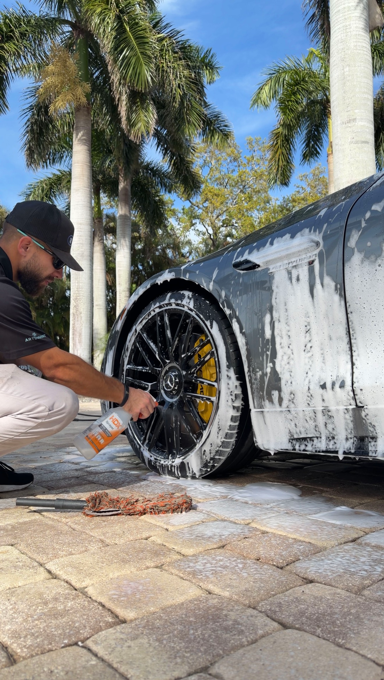 Person cleaning a white sports car wheel with foam outdoors on a sunny driveway.