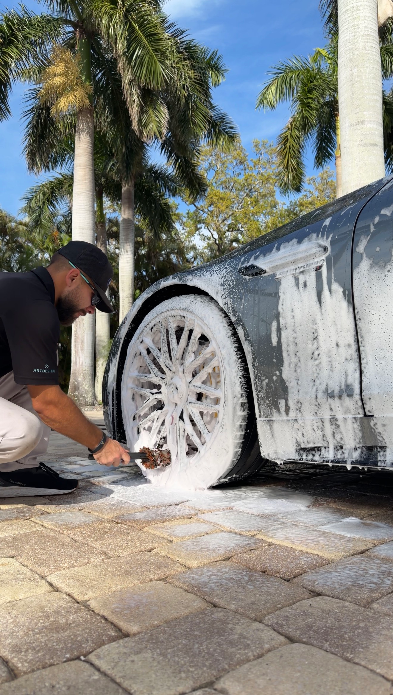 Man washing a white car wheel with foam in a sunny driveway, palm trees in the background