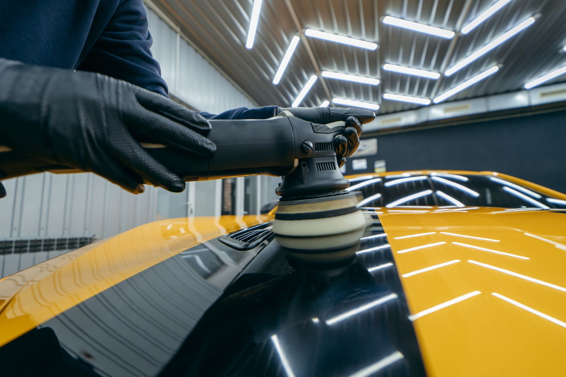 Car detailer polishing a yellow and black car hood under bright workshop lights