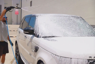 Person pressure-washes a white SUV in a driveway with soap foam covering the windshield.