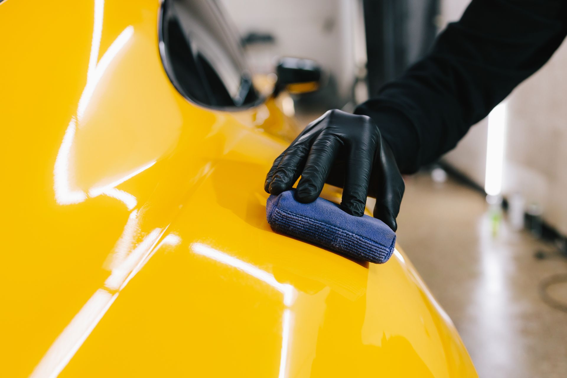Gloved hand polishing a bright yellow car with a blue cloth in a garage