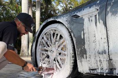 Man washing a white car’s wheel and lower body with soapy water outdoors