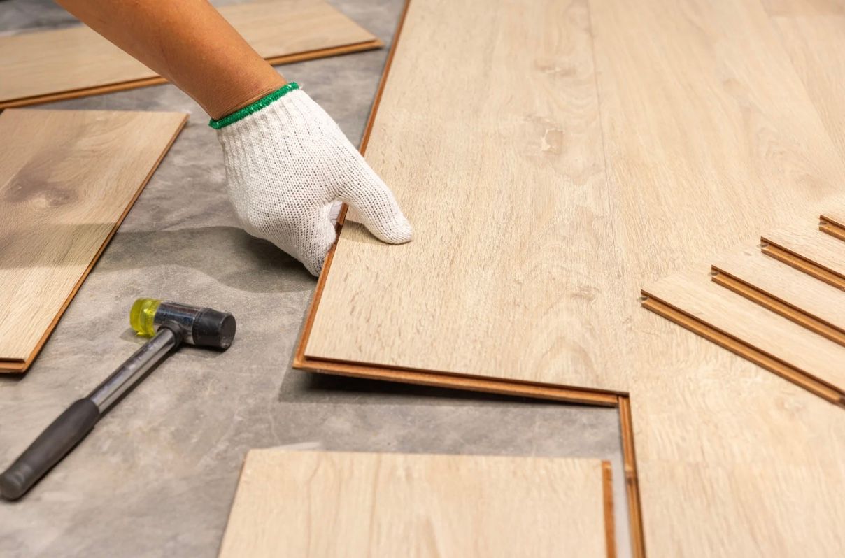 Gloved hand aligning wood flooring planks beside a hammer on a work surface