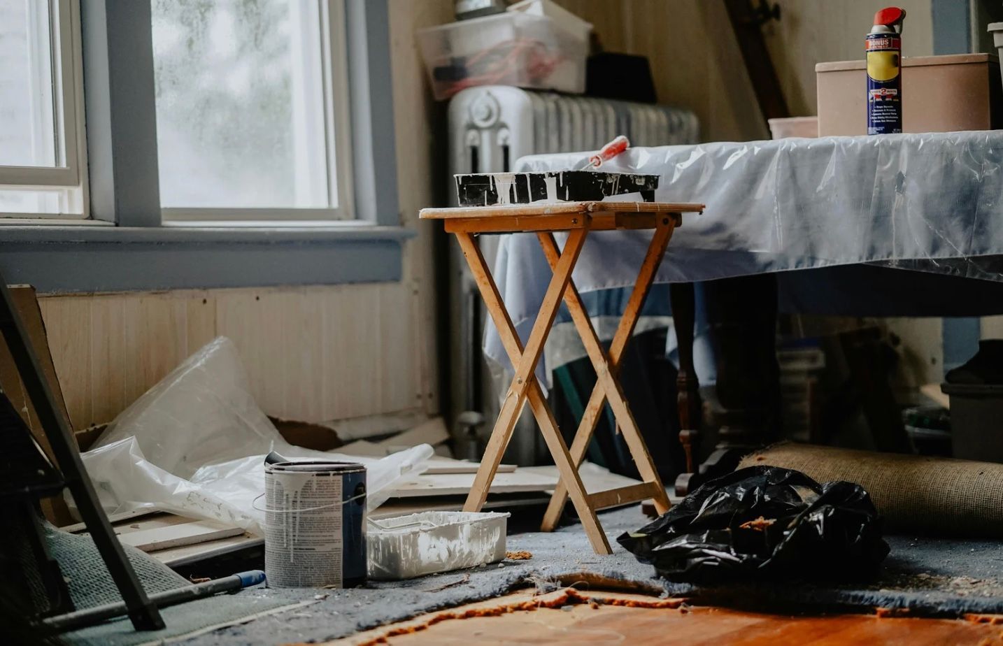 Paint supplies and tools on a messy floor beside a small folding table in a sunlit room