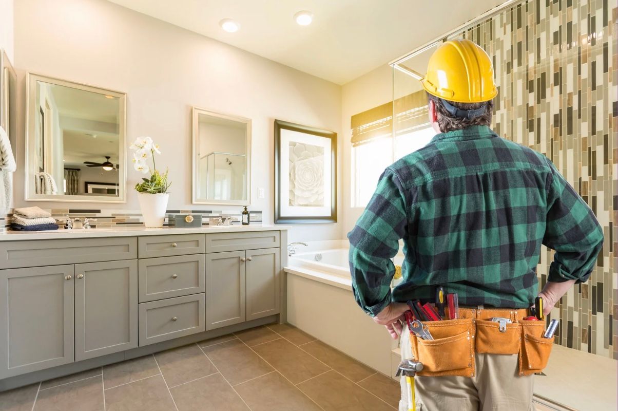 Worker in hard hat stands in a bright bathroom with vanity, mirrors, and a tiled accent wall.