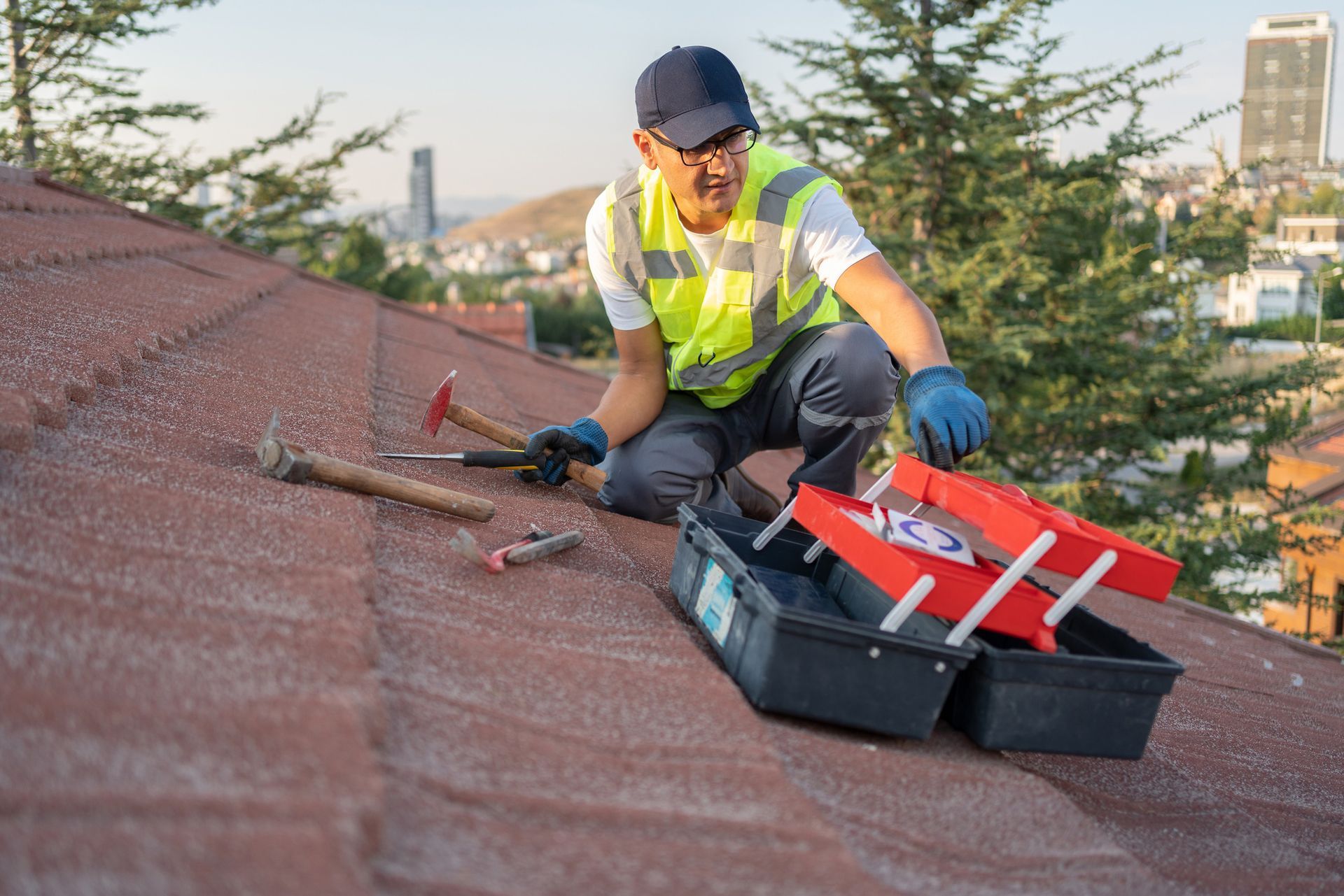 Roofer kneeling on roof, opening toolbox. Wearing reflective vest, cap, and gloves. City in background.