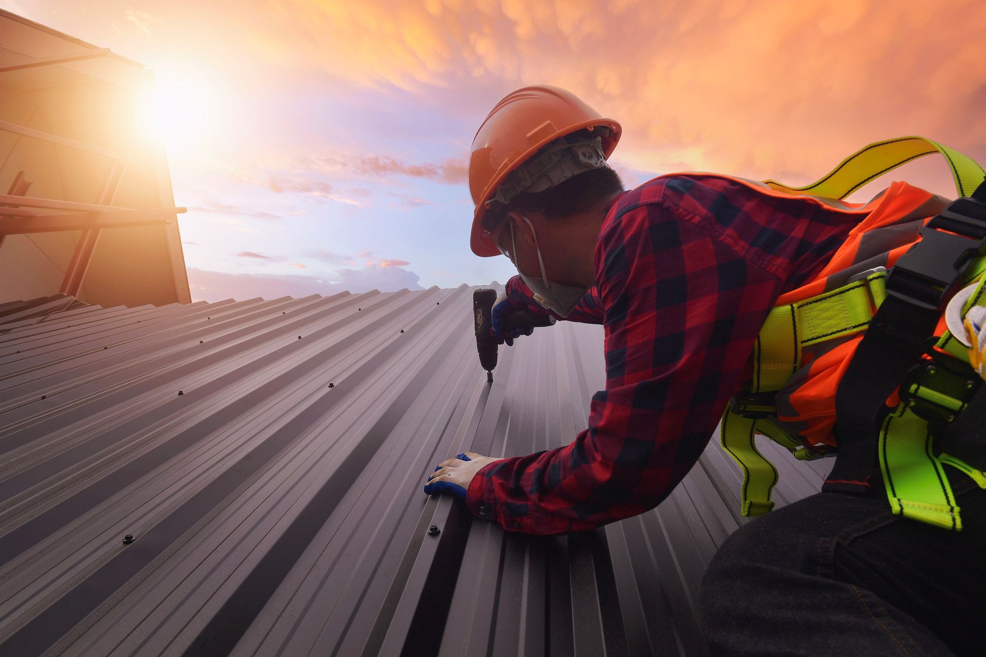 Roofer in safety gear installing metal panels on a roof at sunset.