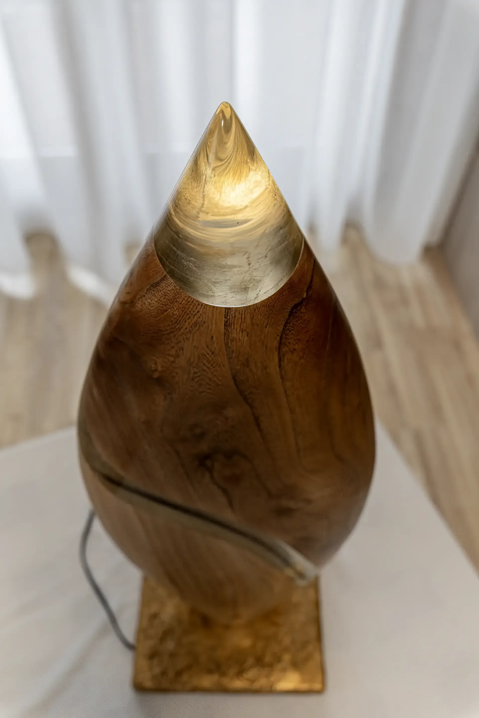 Lampada scultura in resina epossidica trasparente e legno. L'opera ha una forma a goccia  con una sezione principale in legno e un inserto curvilineo in resina. La punta superiore è in resina trasparente dove viene emessa la luce. Opera di Guerino Rusconi.