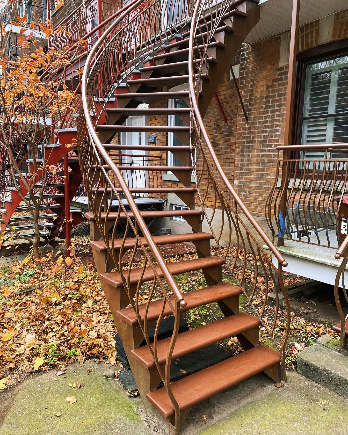 Rust-colored outdoor spiral staircase beside brick building, with fallen leaves and wrought-iron railing.