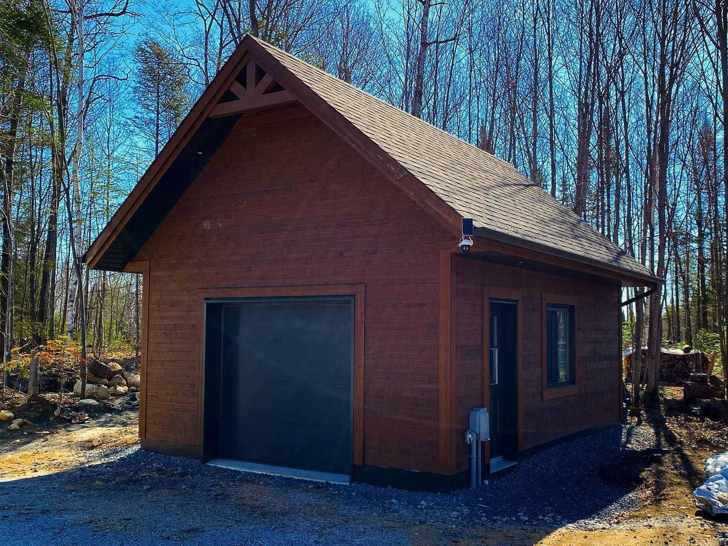 Small wooden shed with a dark garage door and steep roof in a wooded area on a sunny day