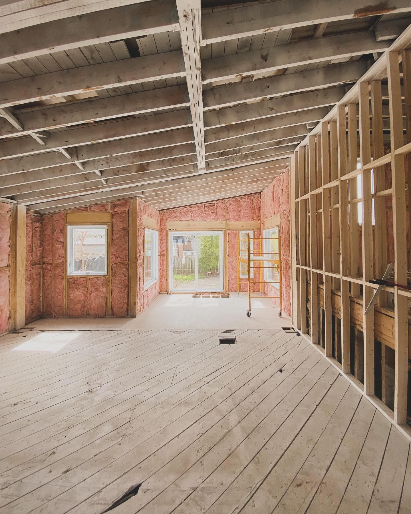 Interior of a house under renovation with exposed framing, unfinished floor, and bright windows at the back.