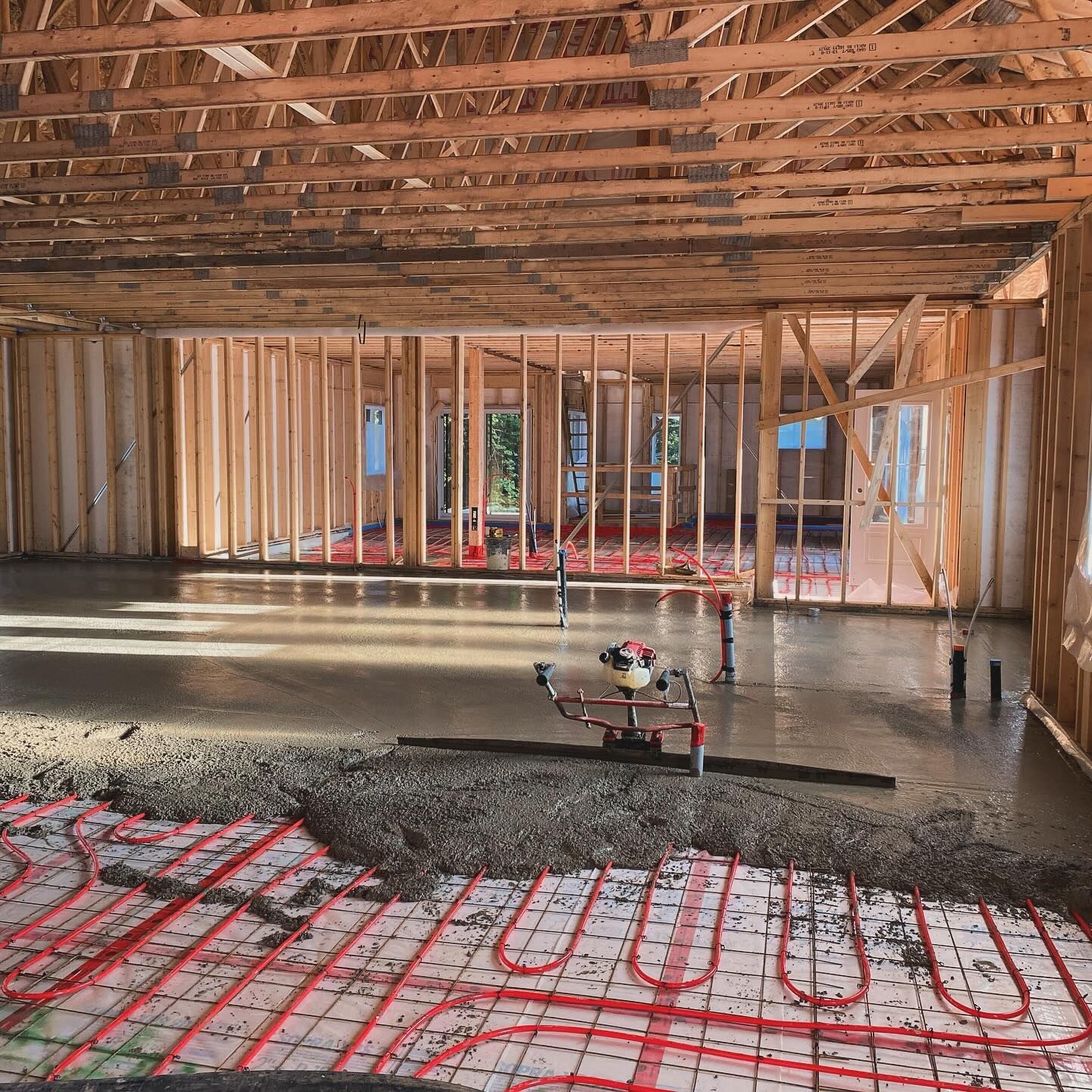 Interior of a house under construction with exposed wood framing, concrete floor, and radiant floor tubing visible