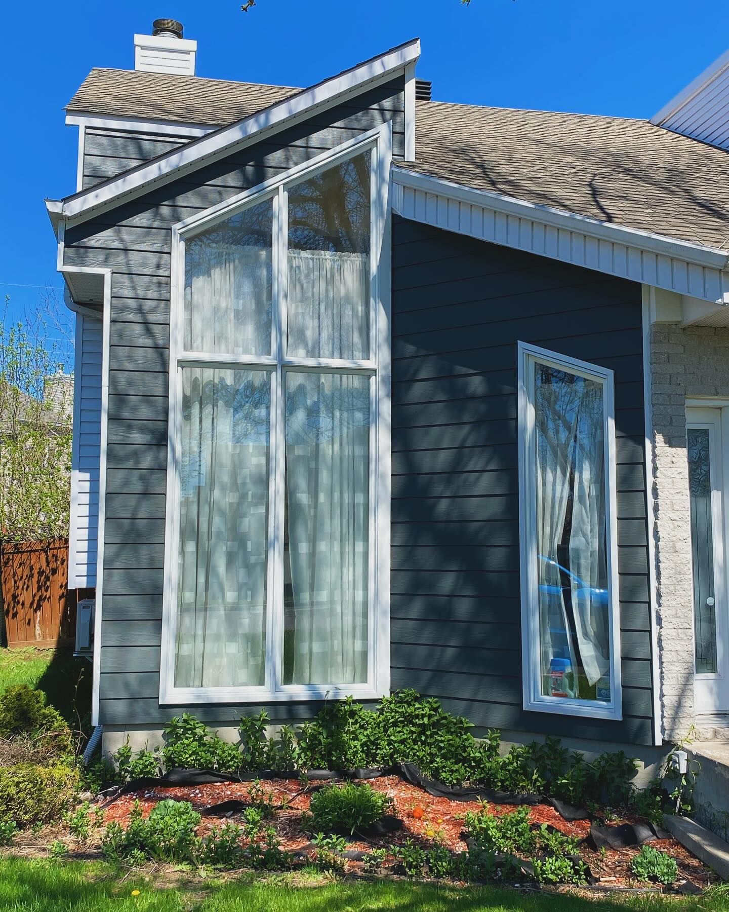 Gray house exterior with tall windows, sloped roof, and landscaped garden in sunlight
