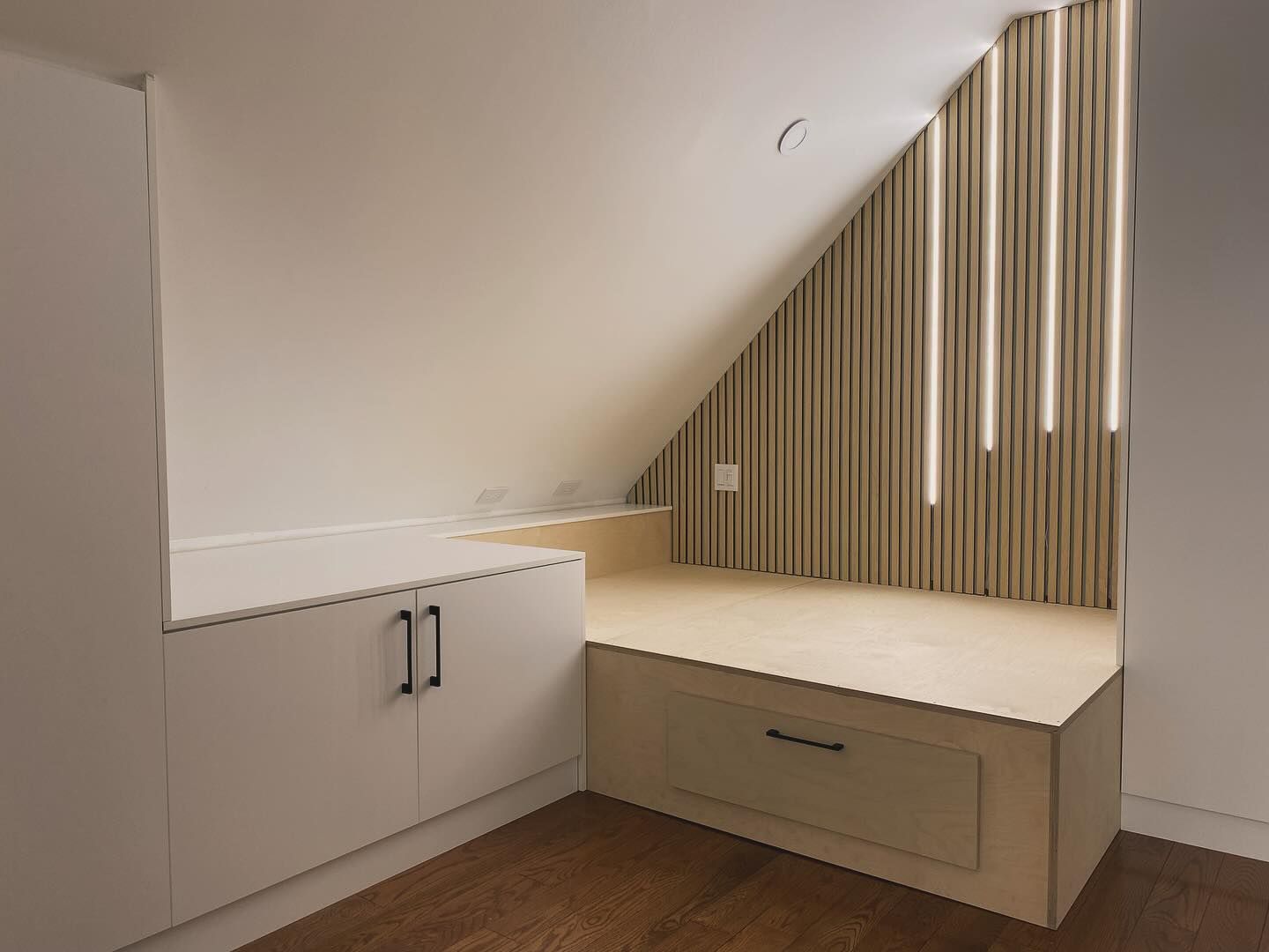 Minimalist attic room with white cabinets, beige bench, and vertical wood slat wall under sloped ceiling