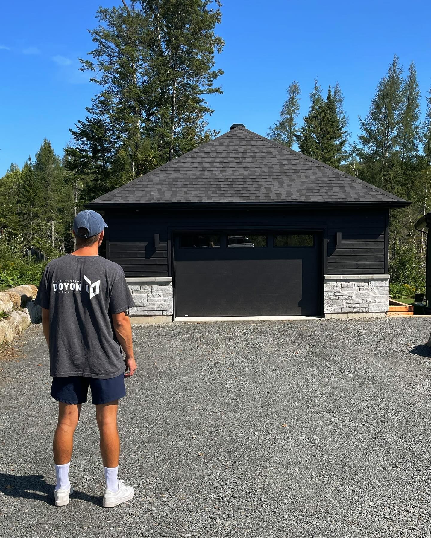 Person standing on gravel driveway facing a dark garage with a shingled roof and trees behind.
