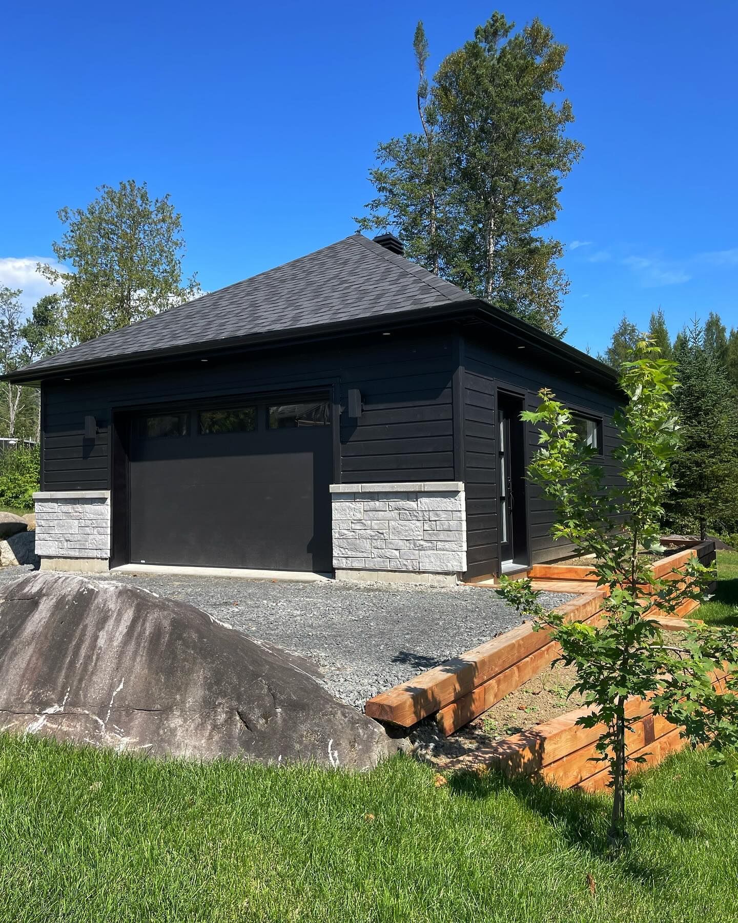 Black detached garage with stone base and dark siding on a sunny wooded property