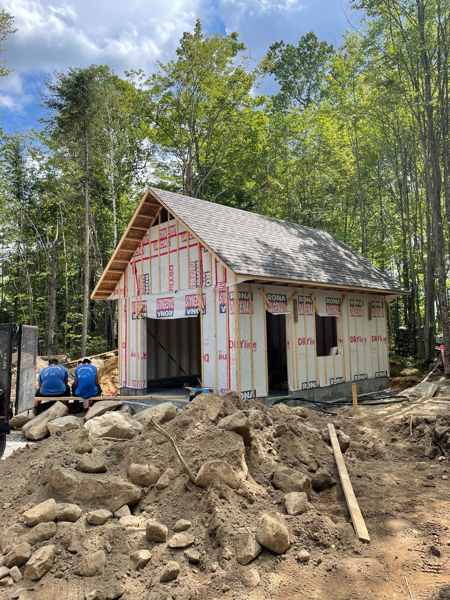 Small house under construction with white siding and red trim, surrounded by dirt and trees.