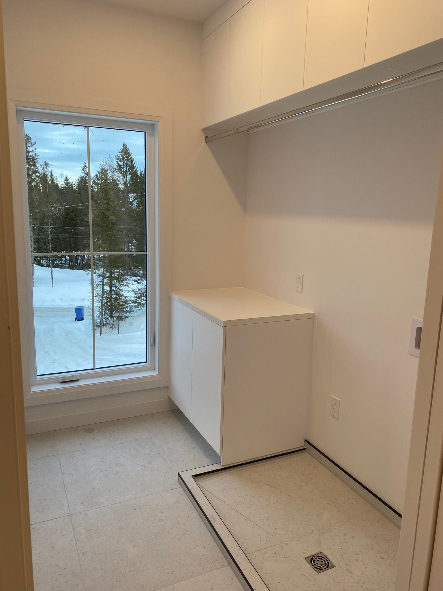 Empty utility room with a window, white cabinet, and a floor drain near a wall-mounted shelf.