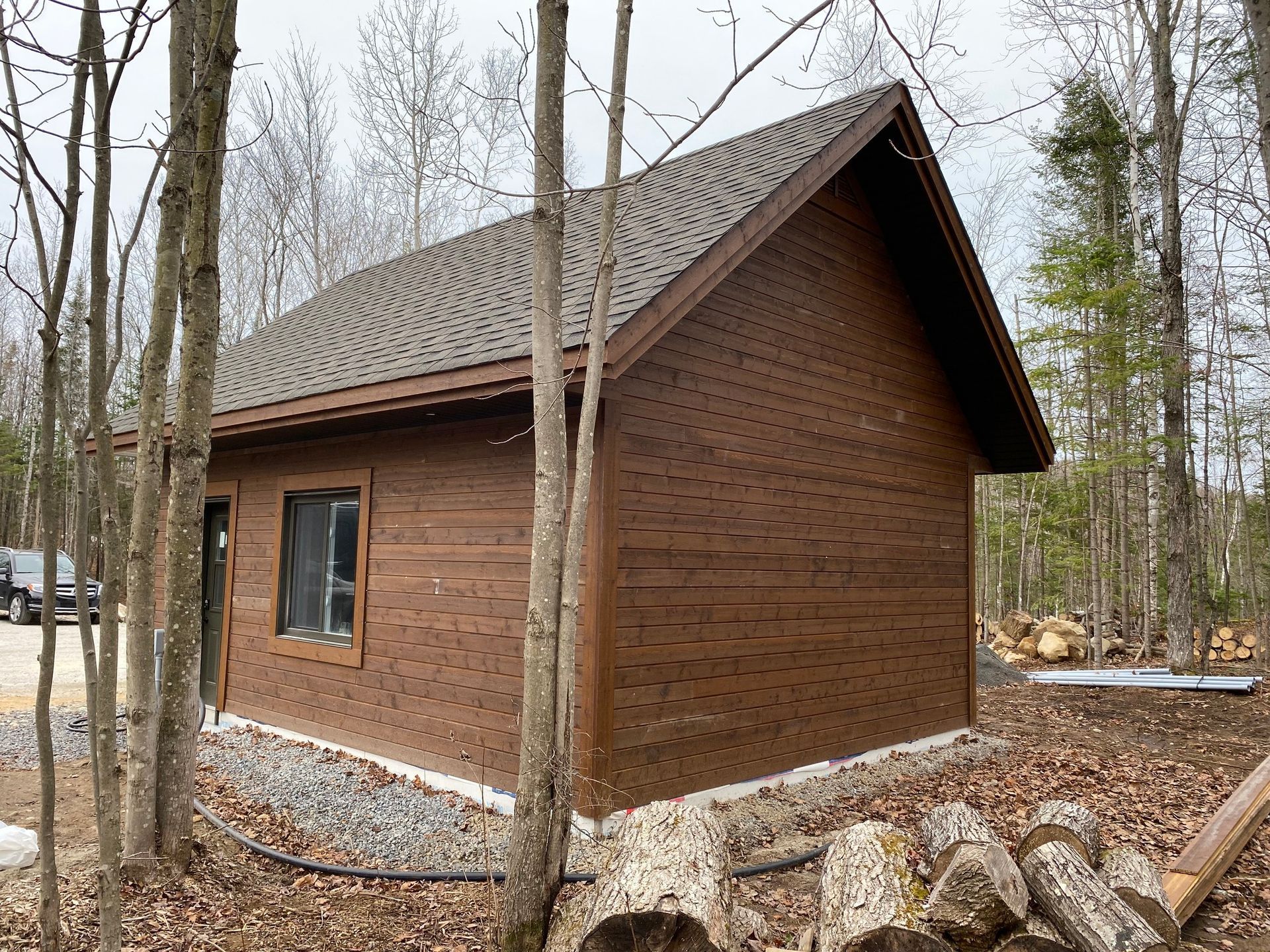 Small red-brown wooden cabin with a dark roof in a leafless forest, surrounded by gravel and rocks
