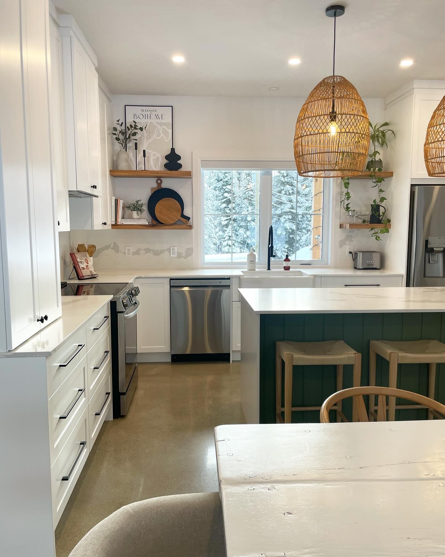 Bright white kitchen with green island, woven pendant lights, and a window overlooking snowy trees.