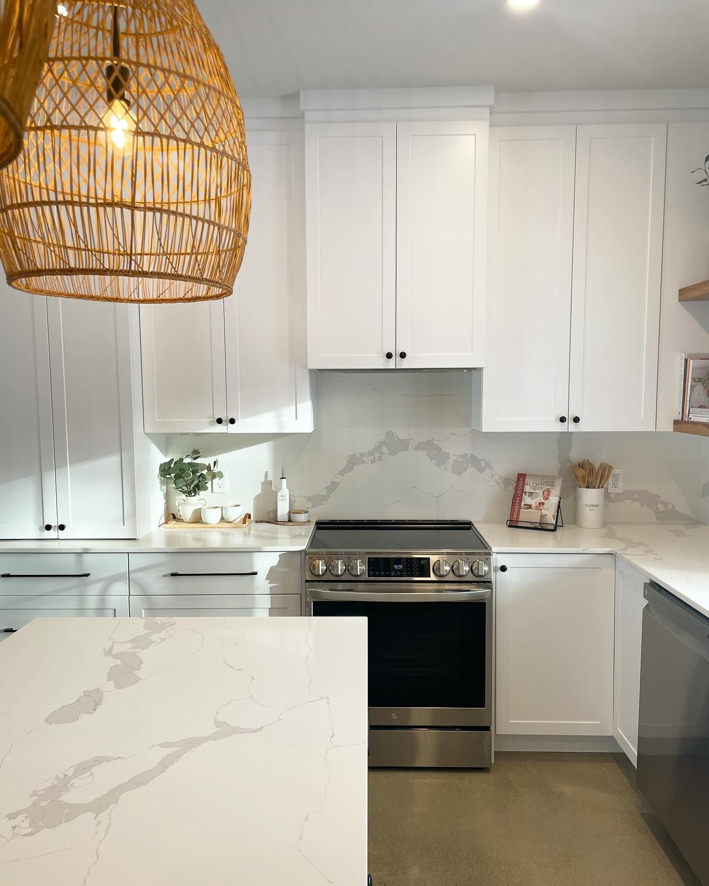 Bright white kitchen with marble counters, stainless stove, dark dishwasher, and woven pendant light