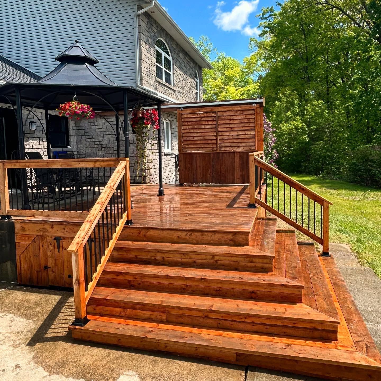 Wooden steps and deck with railing leading to a stone house and gazebo in a sunny yard