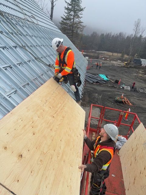 Two workers in safety gear installing plywood on a steep roof at a construction site