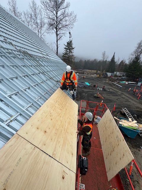 Two workers in safety vests on a sloped roof beside plywood wall panels on a construction site