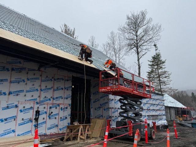 Workers on a red scissor lift repairing a roof on a house wrapped in protective siding