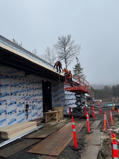 Workers on a red lift installing siding on a house under construction, with orange cones along the wet driveway.