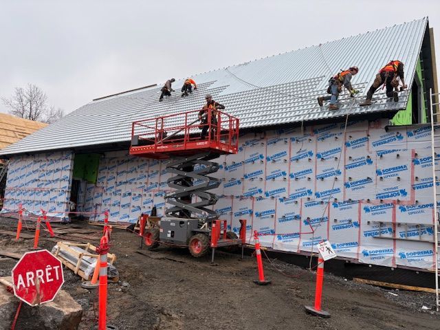 Workers installing a metal roof on a house, with a red scissor lift and safety cones on site.