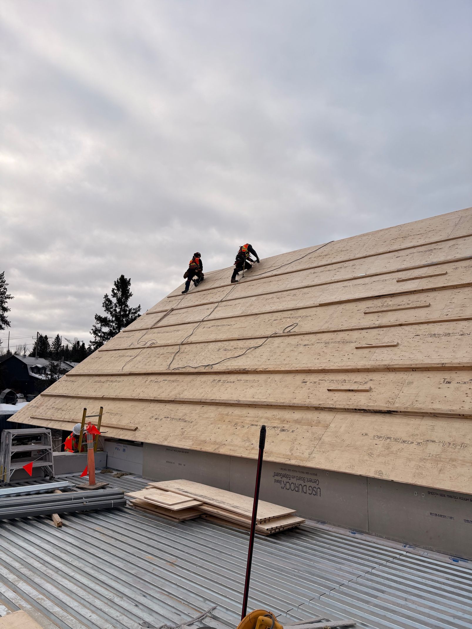 Two roofers installing underlayment on a steep house roof with snow on the ground below.