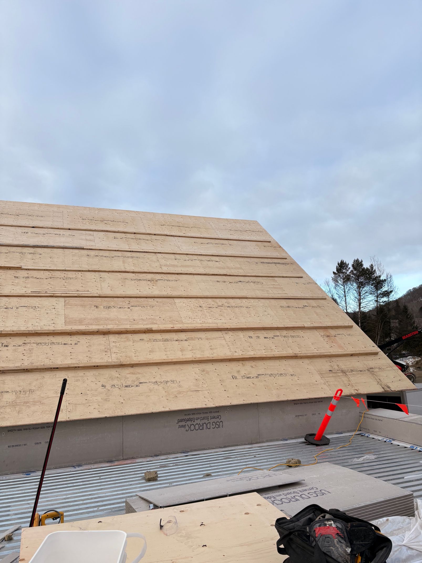 Roof under construction with plywood sheathing, scaffolding, and tools against a cloudy sky