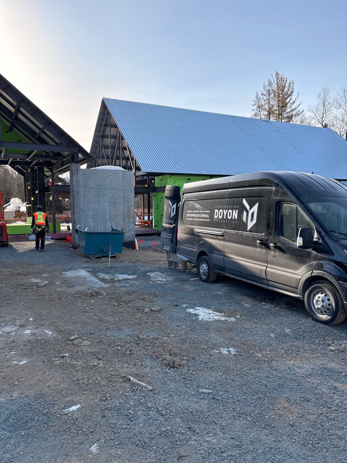 Black van parked on a muddy lot beside a barn and silo under a snowy roofline