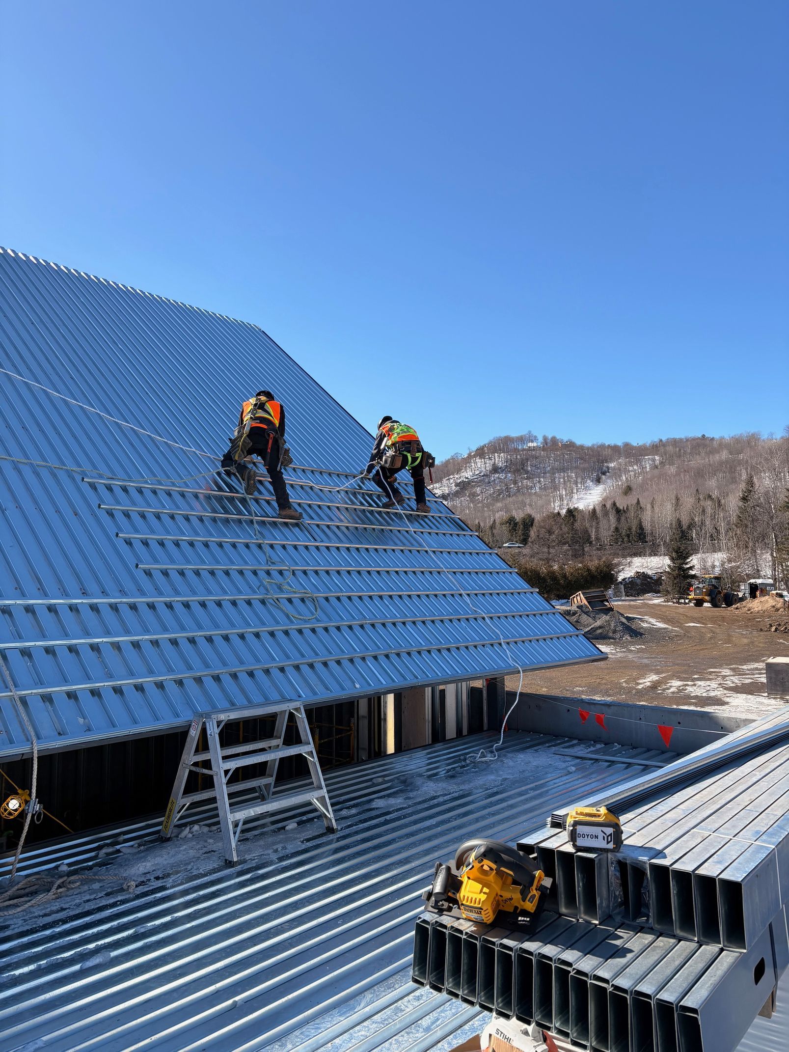 Workers installing blue metal roofing on a steep roof in a snowy mountain setting
