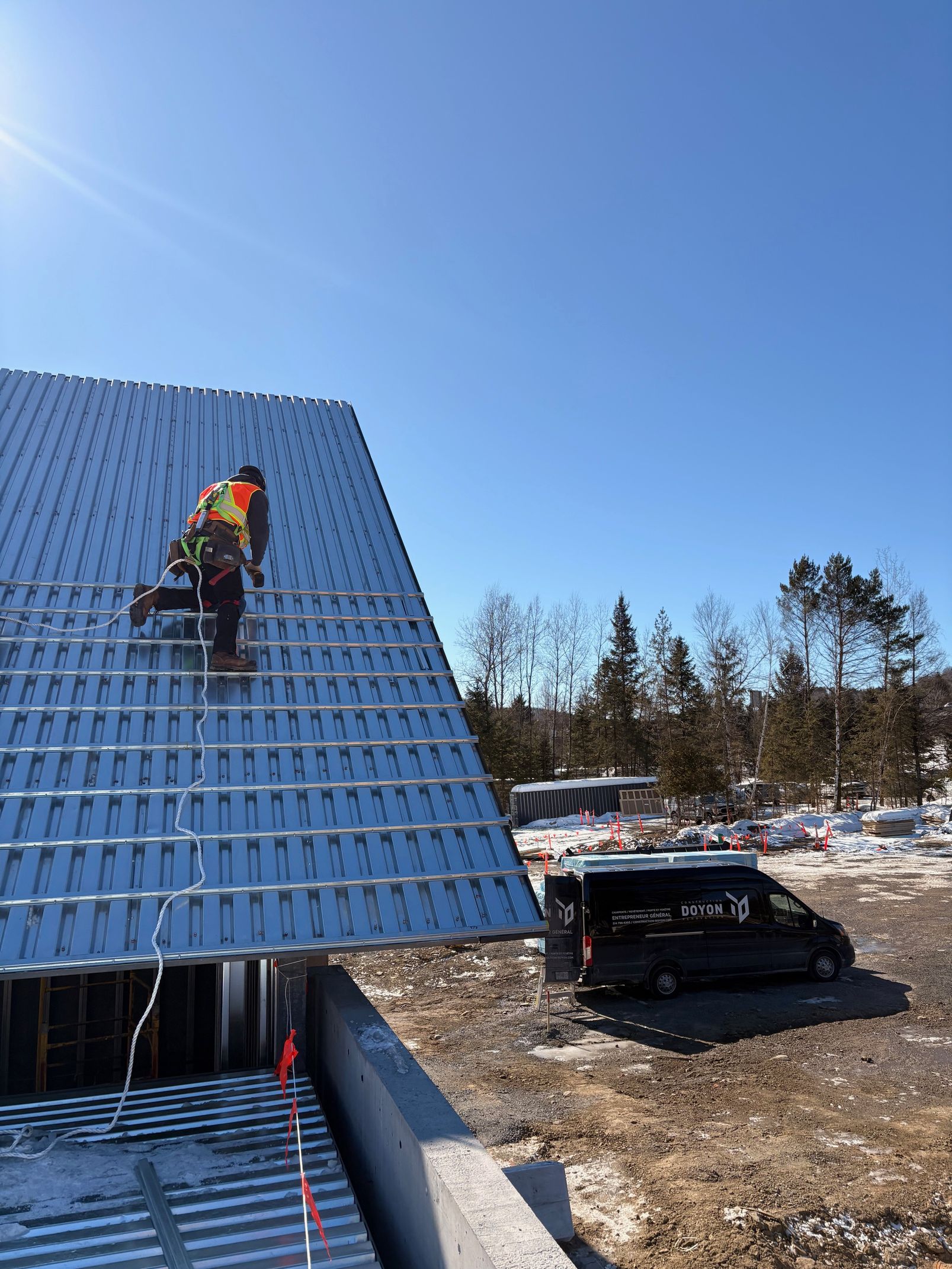 Worker in safety gear climbing a steep blue roof beside a parked truck on a sunny day
