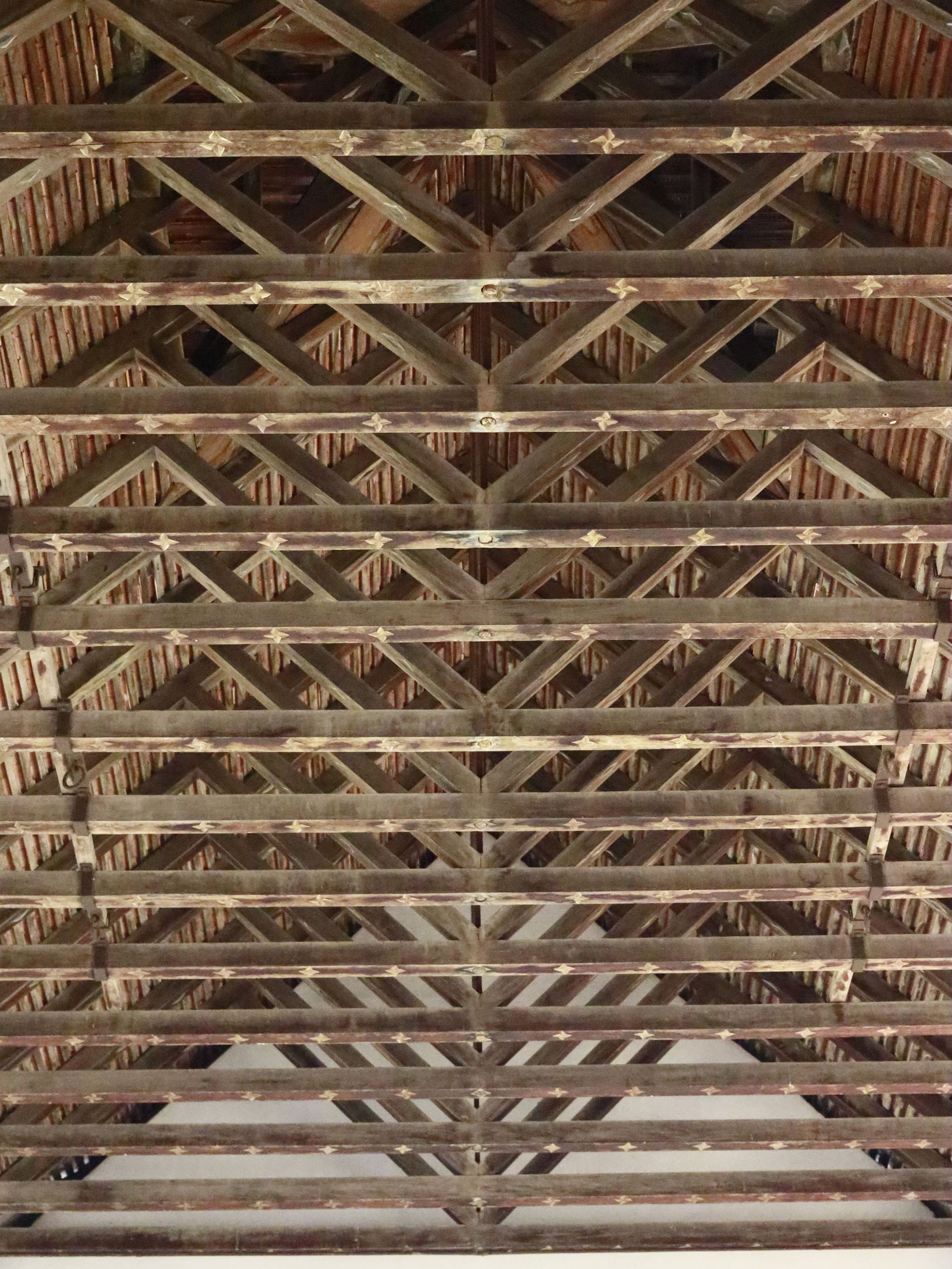 Wooden roof trusses crisscrossing beneath an aged ceiling, viewed from below.