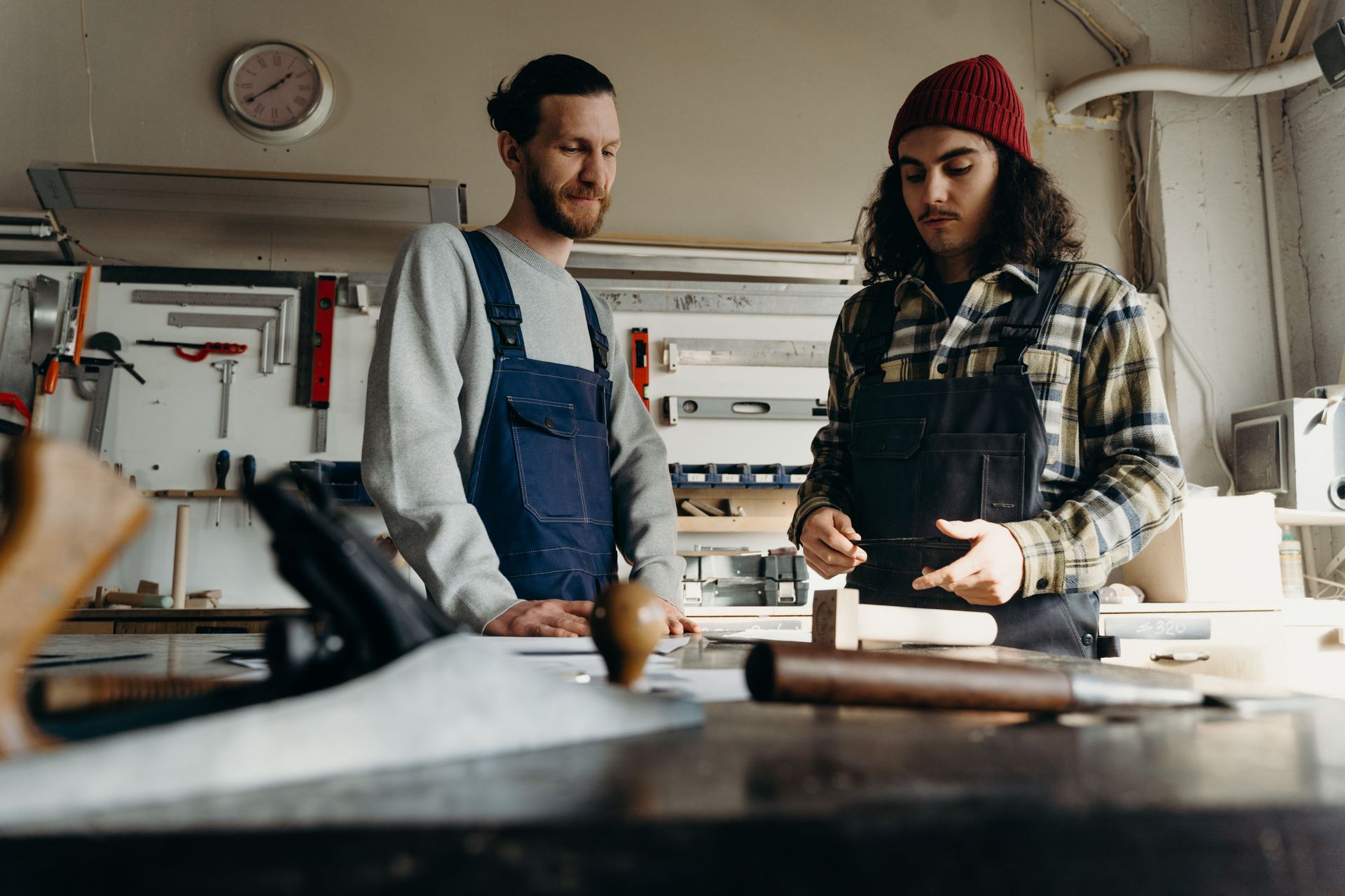 Two people in aprons work at a cluttered workshop table with tools and materials.