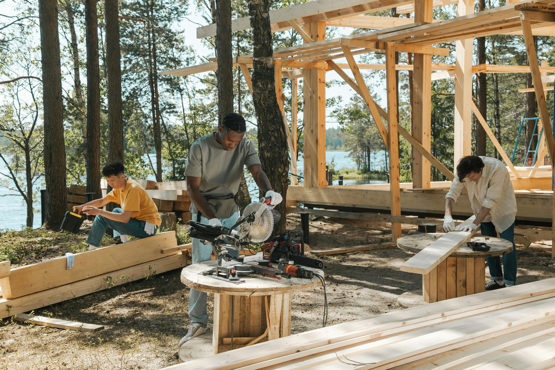 People assembling wooden furniture at a sunny outdoor worksite near trees and a lake.