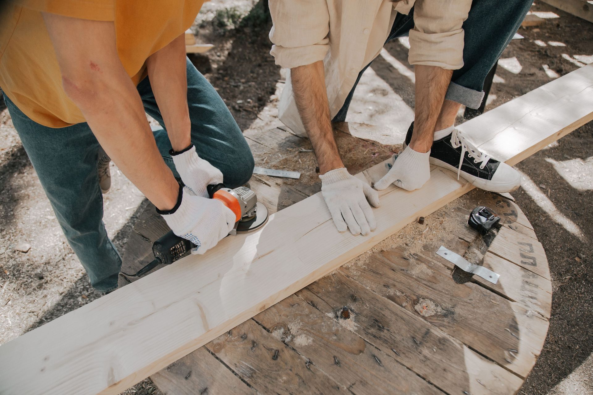 Two people in gloves sanding a white wooden board on a workbench outdoors.