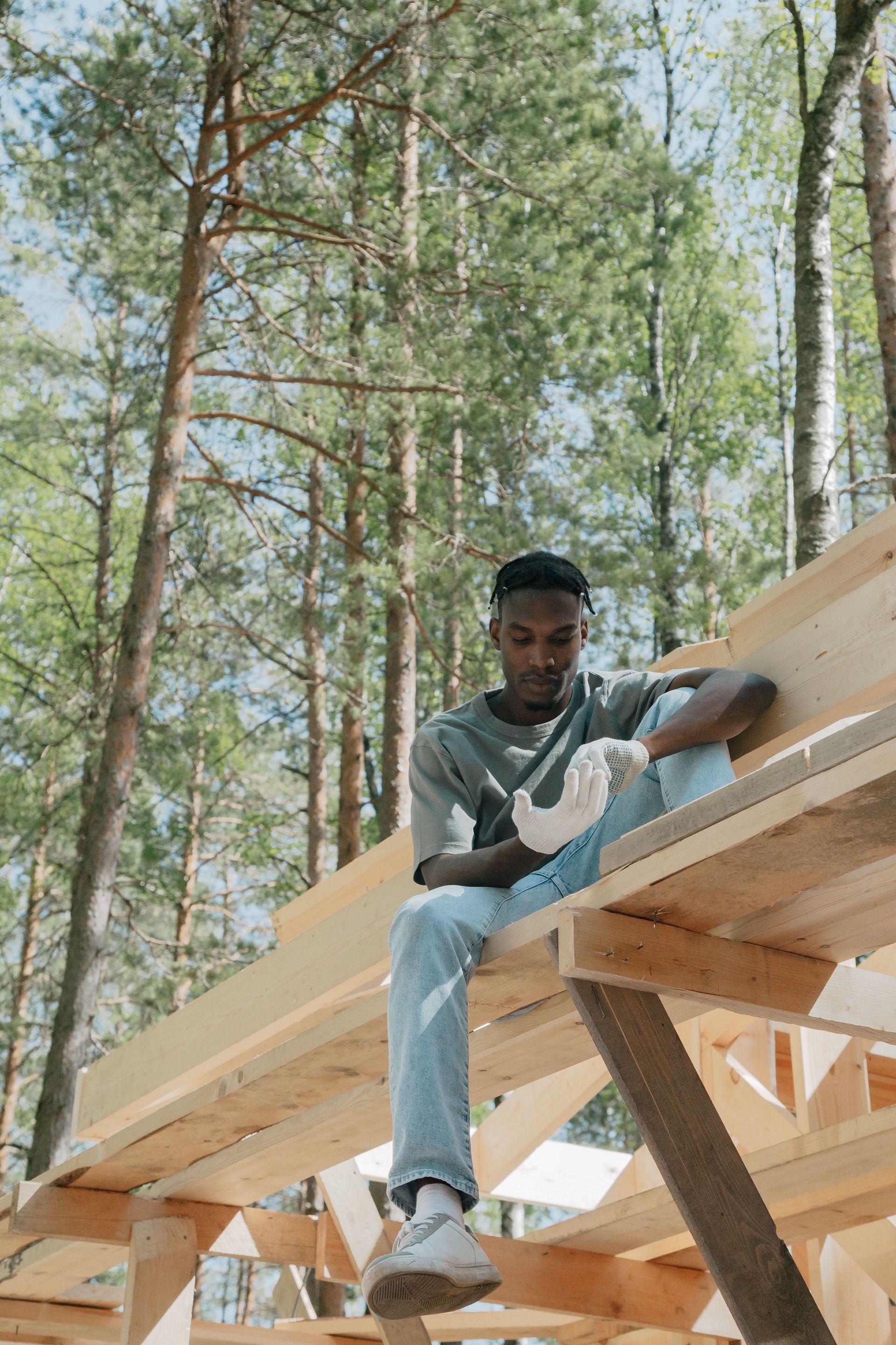 Person sitting on wooden beams at a construction site in a forest, wearing a gray shirt and jeans.