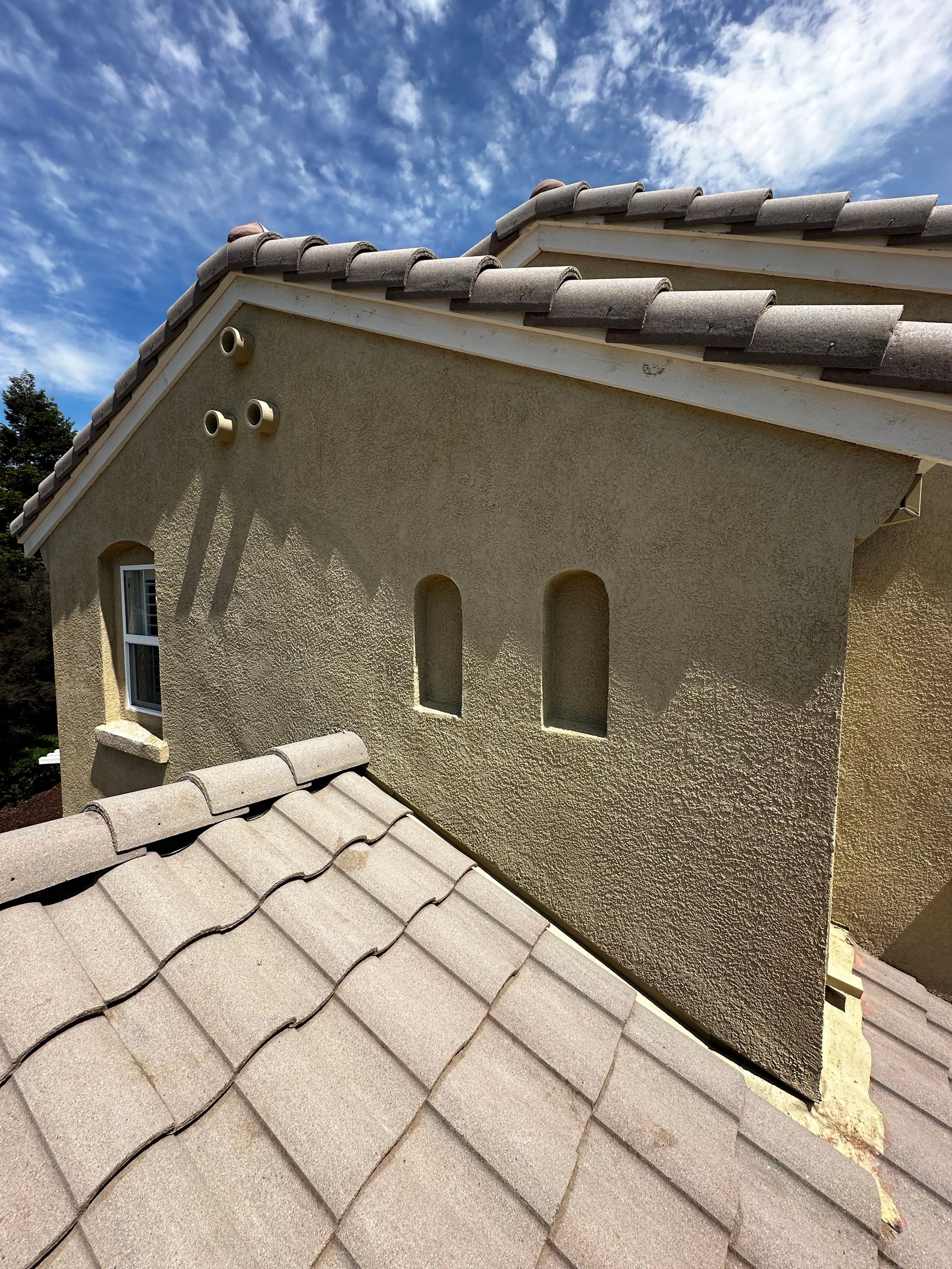 Tan stucco wall of house with tiled roof, arched windows and vents against a blue sky.
