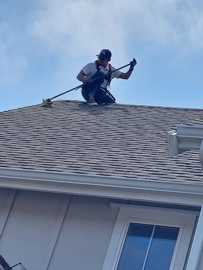 Person on a roof, secured by a harness, cleaning or repairing with a long-handled tool.