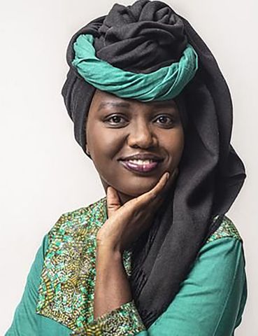 Femme coiffée d'un foulard vert et noir et portant une chemise, la main sur le menton, souriante.