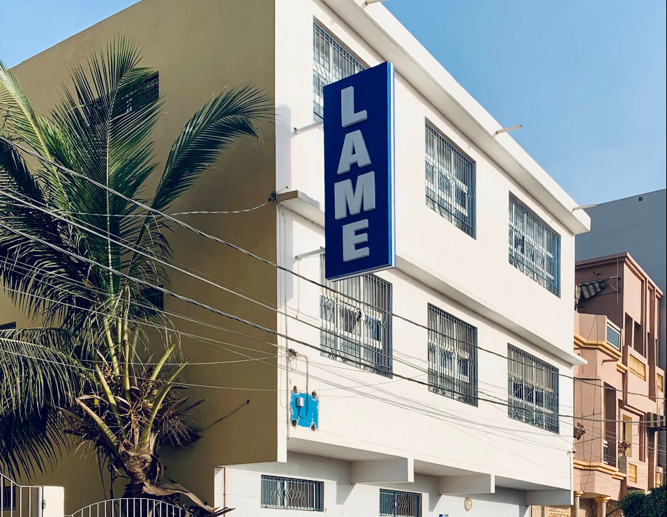 Bâtiment blanc avec un panneau bleu « LAME », un palmier et un ciel bleu en arrière-plan.