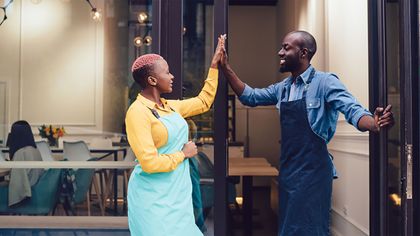 Deux personnes, tablier sur le dos, se tapent dans la main devant l'entrée d'un restaurant. Souriantes.