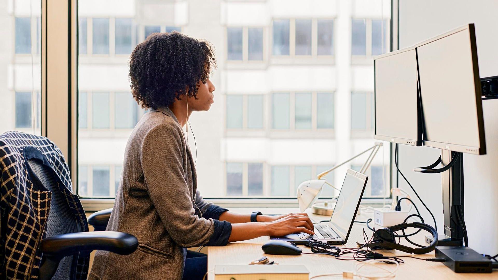 Femme tapant sur un ordinateur portable à un bureau équipé de deux écrans, dans un environnement de bureau.
