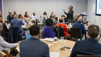 Une femme fait un geste en présentant un exposé à un groupe diversifié assis à des tables dans une salle de conférence.